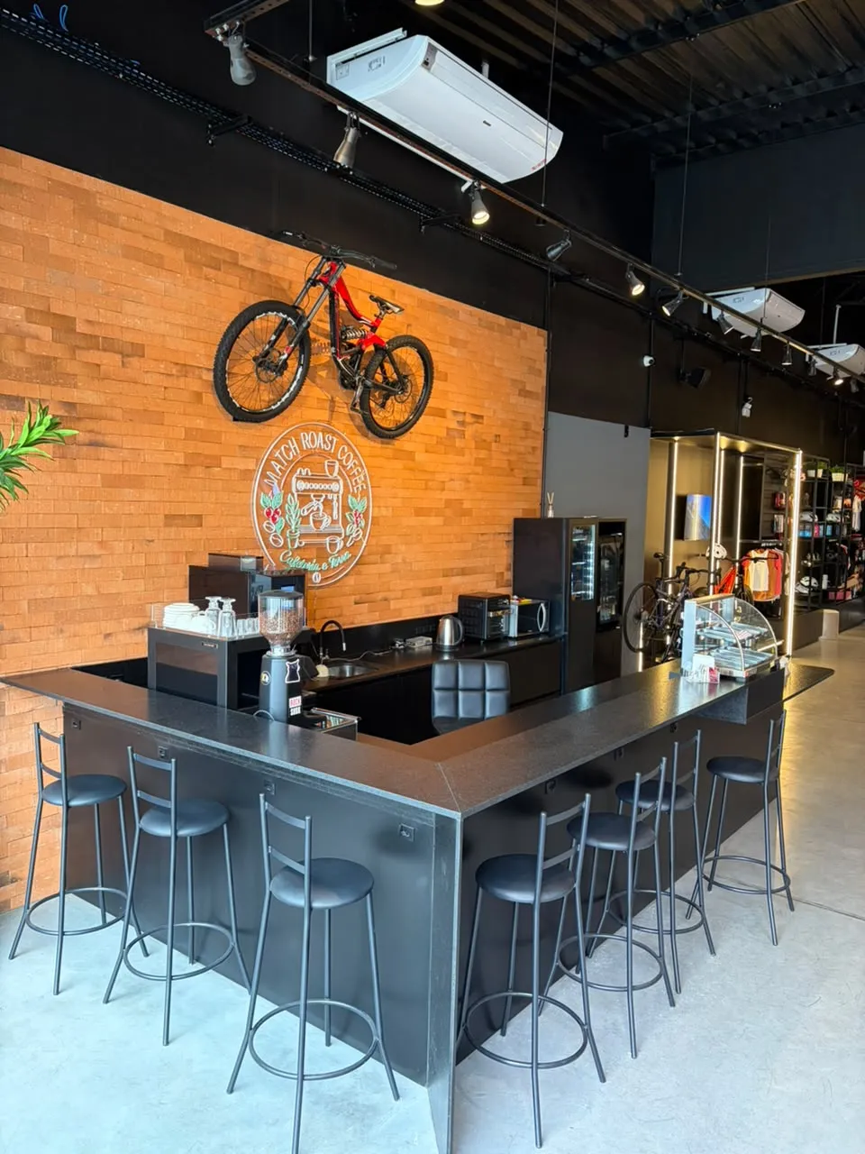 Cafeteria dentro da SB Place, com balcão de granito preto, parede de tijolos e uma bike na parede